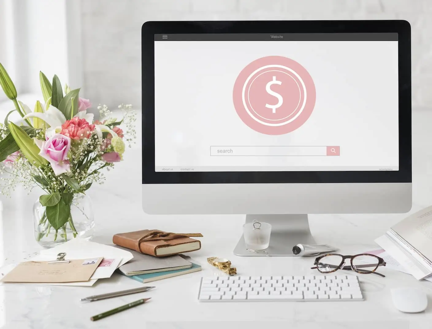 A modern and tidy workspace with a large desktop monitor displaying a website's homepage, prominently featuring a pink dollar sign icon in the center to denote pricing. The desk is adorned with a bouquet of fresh flowers, a closed notebook tied with a strap, personal stationery, a pair of glasses, and a cup, suggesting a blend of professional web design work and personal touch.